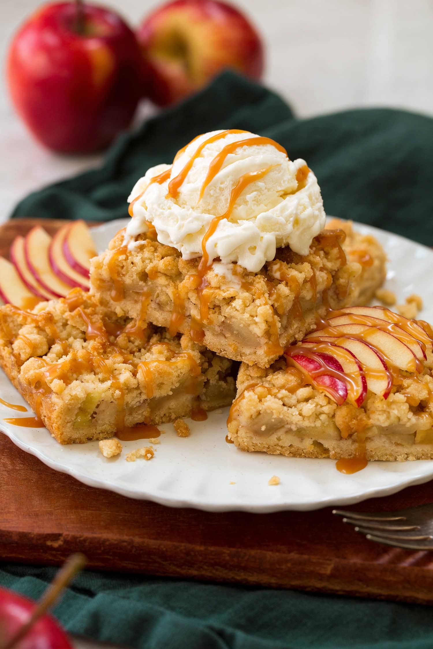 Three apple pie crumb bars stacked on a plate with a scoop of vanilla ice cream on top and drizzled with caramel sauce.