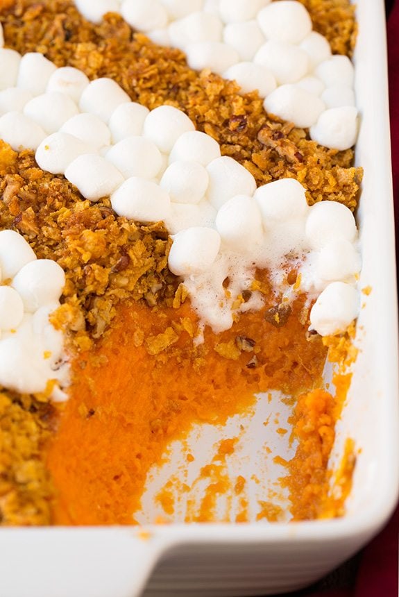 Sweet Potato in a white casserole dish. One scoop is removed to show sweet potato layer. It's topped with rows of crushed cornflakes and pecans then alternating rows of mini marshmallows.