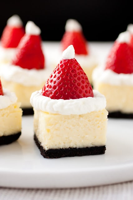 Santa Hat Cheesecake Bites - cheesecake squares layered with oreo crust, white chocolate cheesecake filling and topped with strawberries, whipped cream