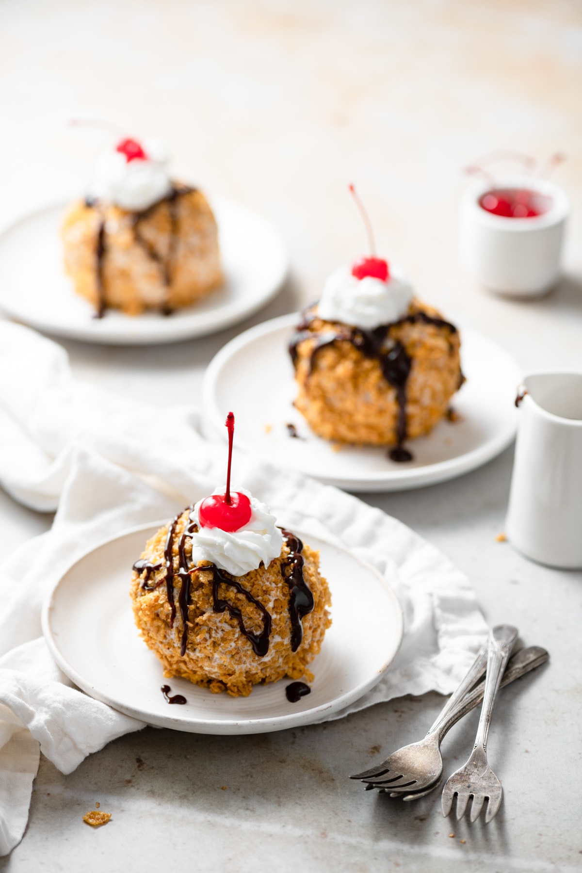 Three individual servings of fried ice cream coated in cinnamon cornflake topping, drizzled with chocolate sauce and garnished with a cherry.