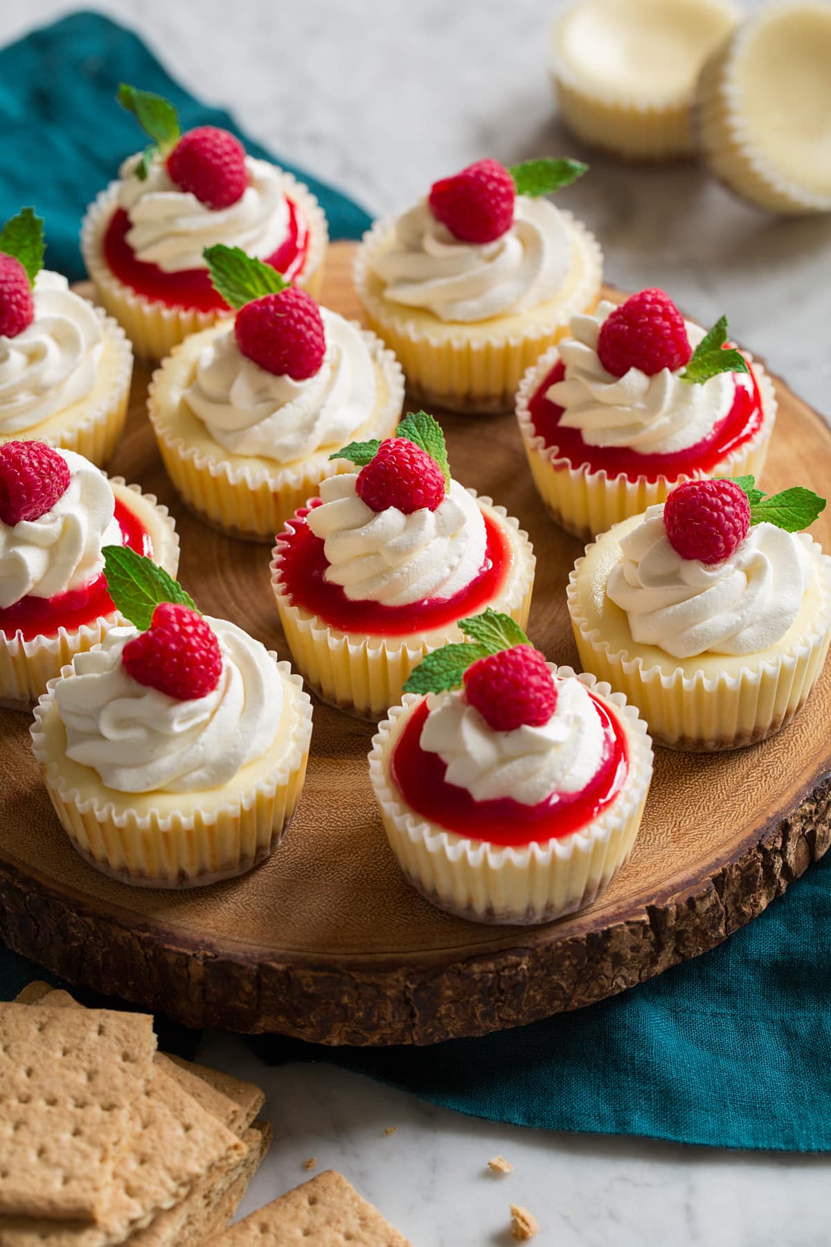 Mini cheesecakes topped with raspberry sauce and whipped cream and mint. Shown sitting on a wooden serving stand.