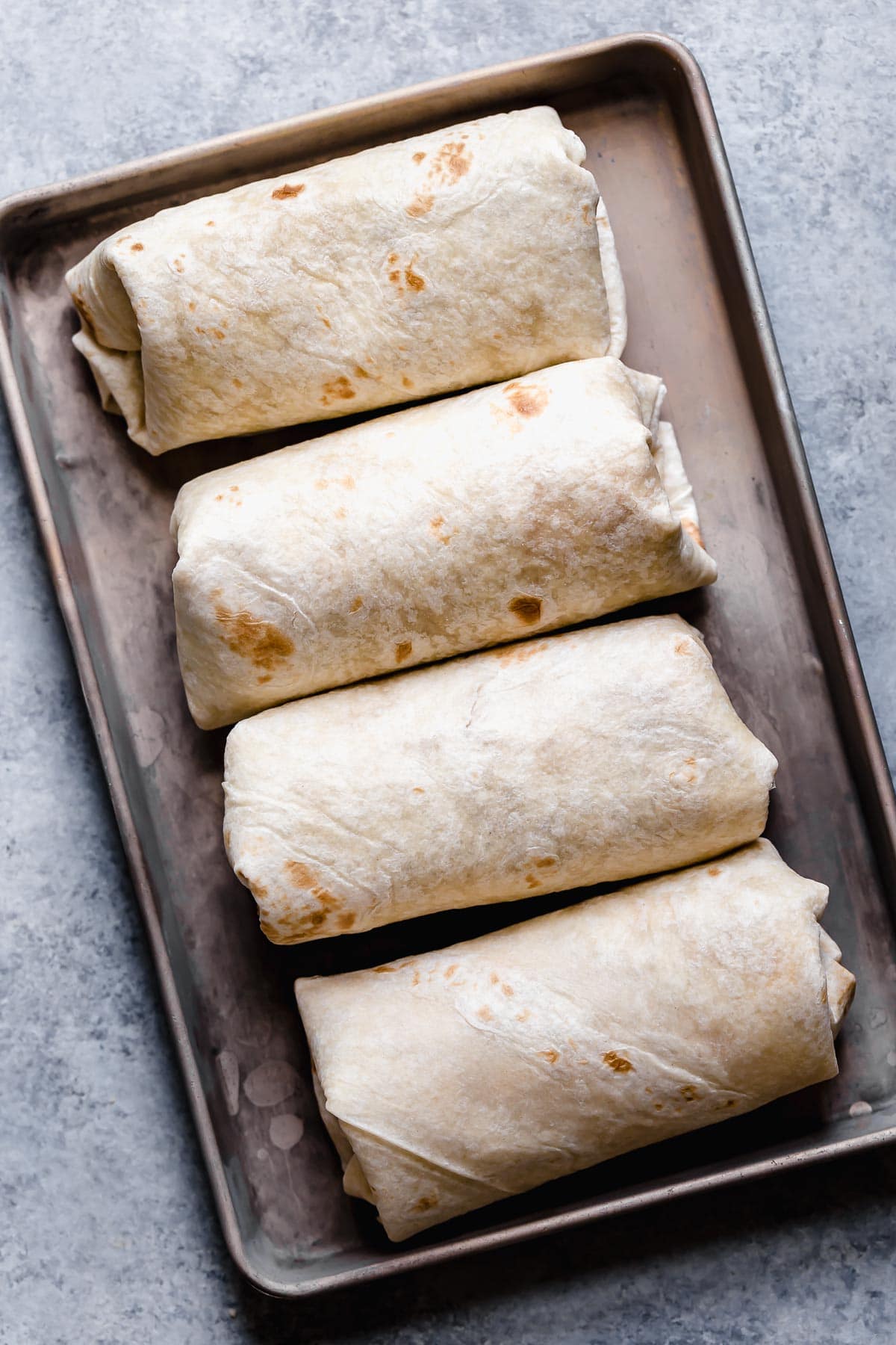 Chicken Chimichangas - one of the ultimate comfort foods! Perfectly hearty and layered with delicious ingredients like seasoned chicken, rich refried beans and shredded Mexican cheese. They're perfectly crisp and oh so satisfying! #chimichanga #chicken #mexican #recipe #refriedbeans #dinnerideas Four assembled homemade chimichangas in a row before frying.