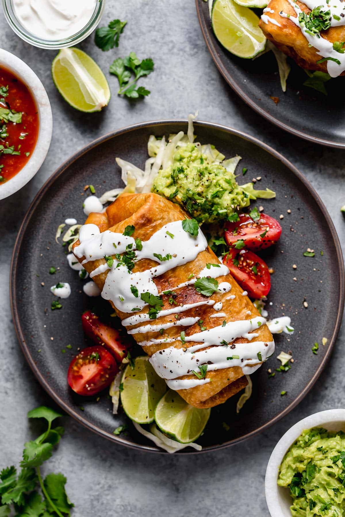 Chicken Chimichangas - one of the ultimate comfort foods! Perfectly hearty and layered with delicious ingredients like seasoned chicken, rich refried beans and shredded Mexican cheese. They're perfectly crisp and oh so satisfying! #chimichanga #chicken #mexican #recipe #refriedbeans #dinnerideas Chicken Chimichanga on a dark plate garnished with guacamole, sour cream, tomatoes and cilantro.