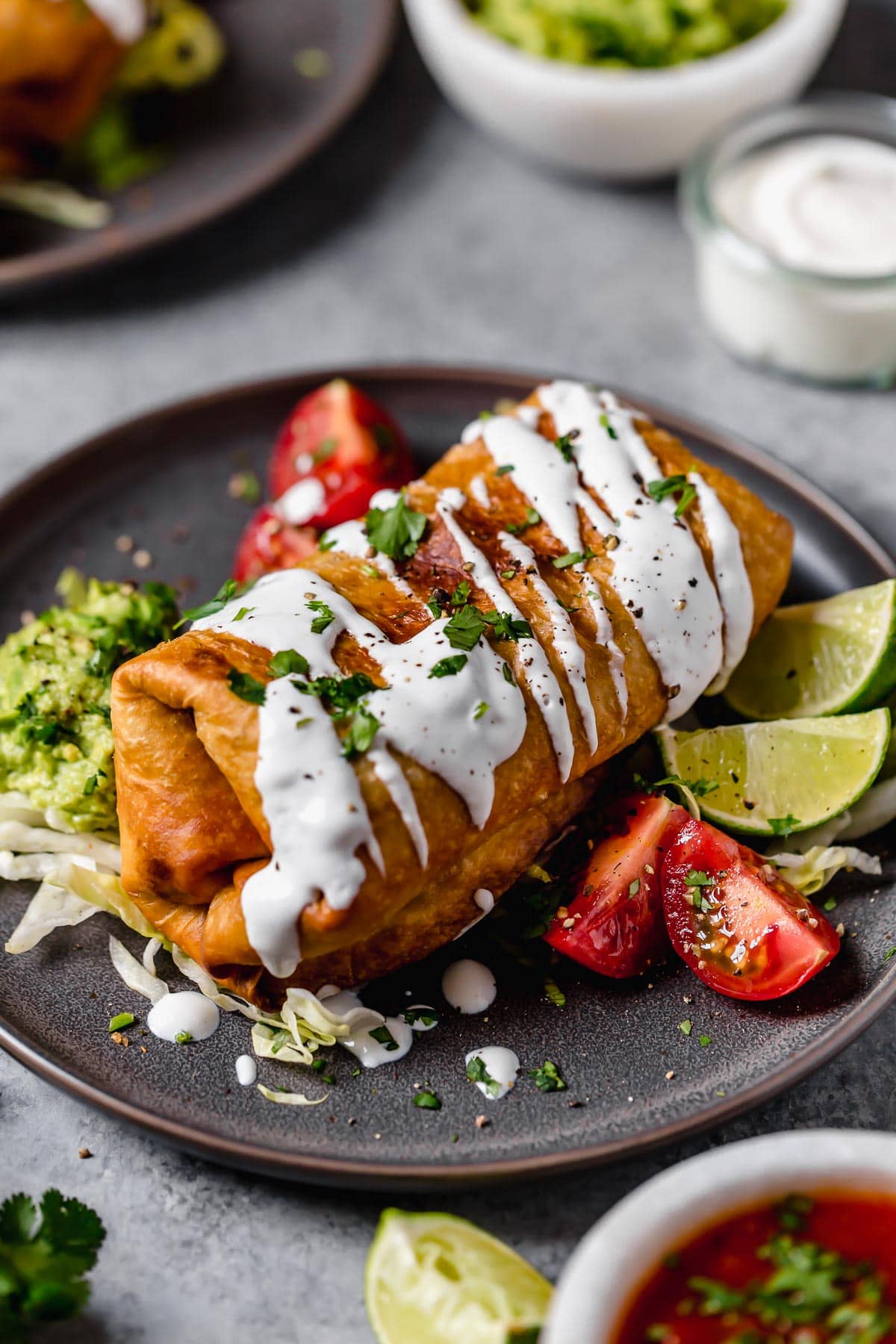 Homemade Chicken Chimichanga on a plate. It's drizzled with sour cream, garnished with cilantro and served with tomatoes and guacamole on the side.