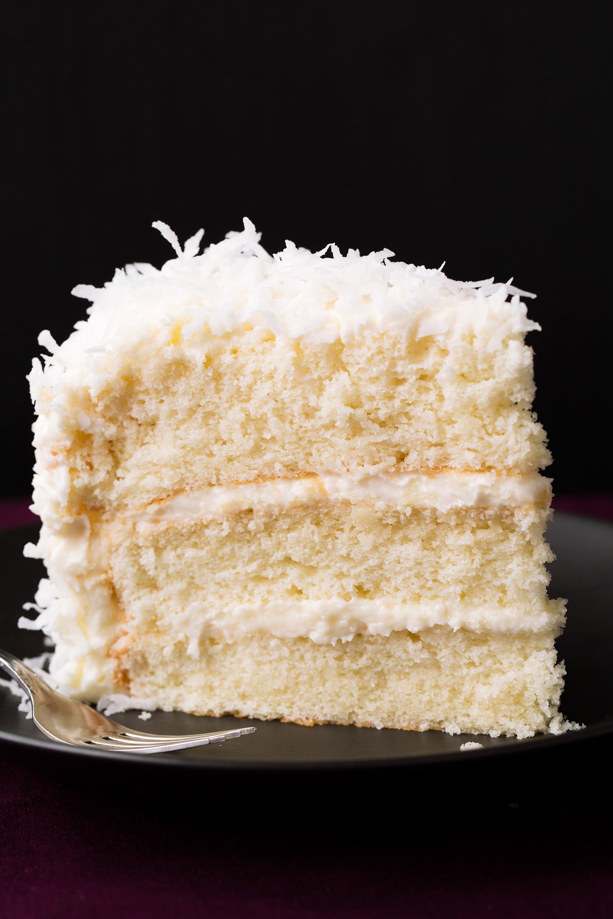 Slice of three layered homemade coconut cake on a black plate with a black background.