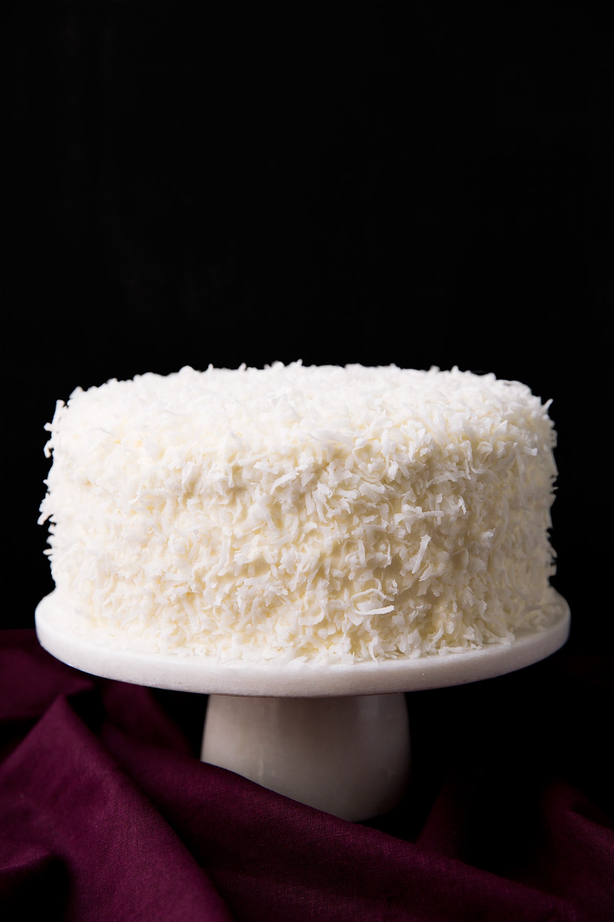 Whole made from scratch coconut cake garnished with lots of coconut and set over a white marble cake stand with a black ground and a maroon tablecloth underneath.