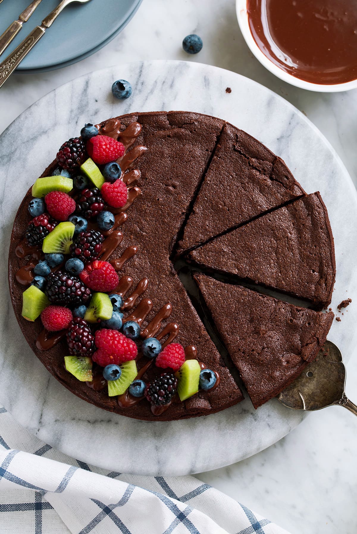Overhead image of flourless chocolate cake on a marble platter. Cake is topped with chocolate ganache drizzle and fresh fruit on the side.
