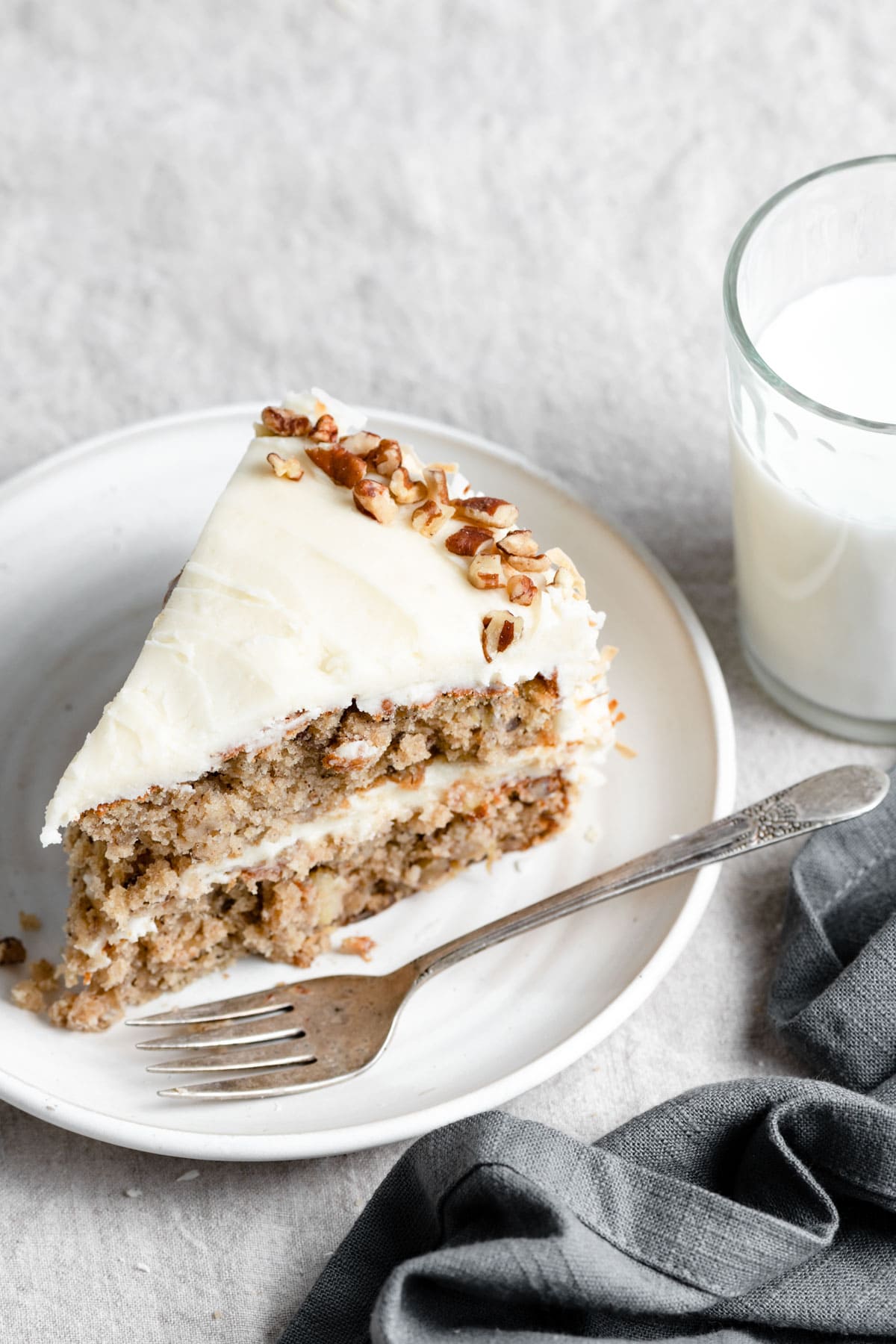 Slice of Hummingbird Cake on a white dessert plate with a fork. Next to they plate there is a dark grey napkin and glass of milk in the background.