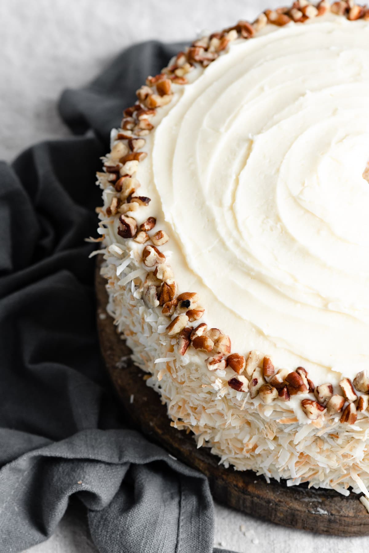 Close up image of whole hummingbird cake showing cream cheese frosting, coconut decorated edges and a ring of pecans on top.