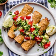 Taquitos on a white plate topped with sour cream, tomatoes, avocados and cilantro. Lime wedges are set of to the side and plate is set on a blue striped napkin.