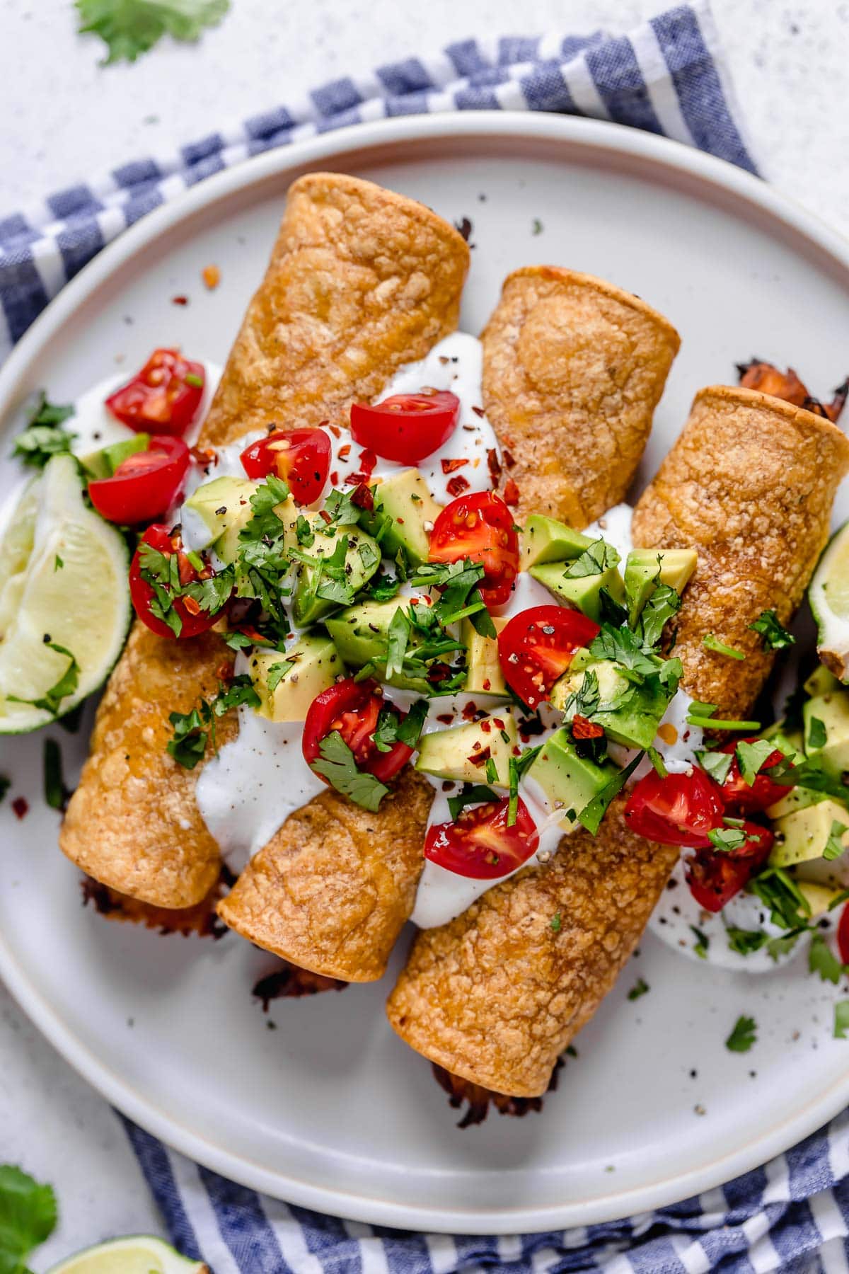 Baked Chicken Taquitos Overhead image of chicken taquitos on a white serving plate topped with sour cream, tomatoes, avocados and cilantro.