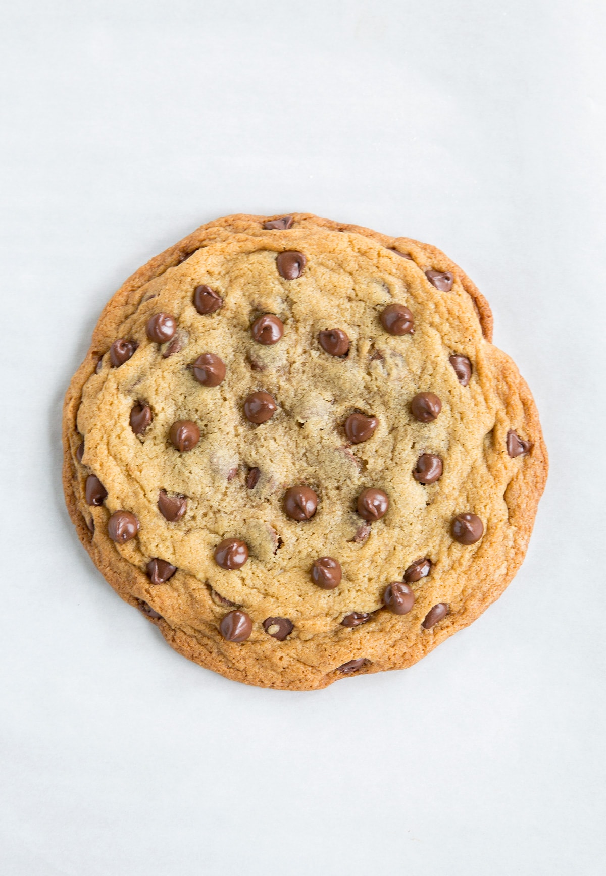 One big chocolate chip cookie shown sitting on white parchment paper.