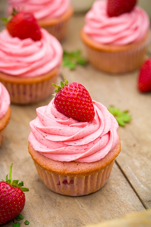 These Strawberry Cupcakes are made with chunks of fresh strawberries in the cake batter and are topped with a luscious pink Strawberry Buttercream Frosting. Bursting with flavor, these cupcakes would go down a treat at any celebration! #cookingclassy #cupcakes #strawberry #buttercream #cakes Strawberry Cupcakes with Strawberry Buttercream Frosting sitting on a wooden surface