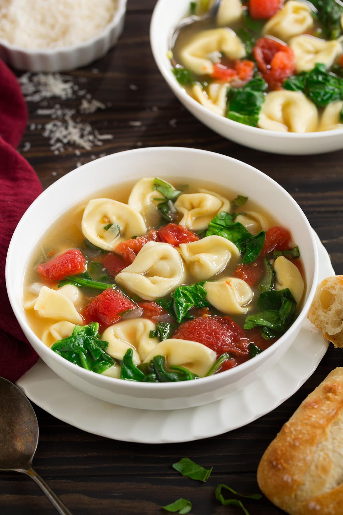Tortellini Soup with Spinach Tomato and Garlic in a white serving bowl set over a ruffled white plate, sitting on a dark wooden tabletop with a side of bread.
