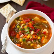 Single serving of vegetable soup in a white bowl set over a white plate. Set on a dark wooden table with a red napkin to the side.