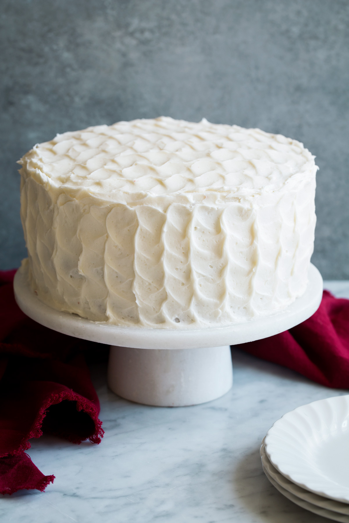Whole red velvet cake covered in cream cheese frosting with a ruffled design. Cake is sitting on a marble cake stand.