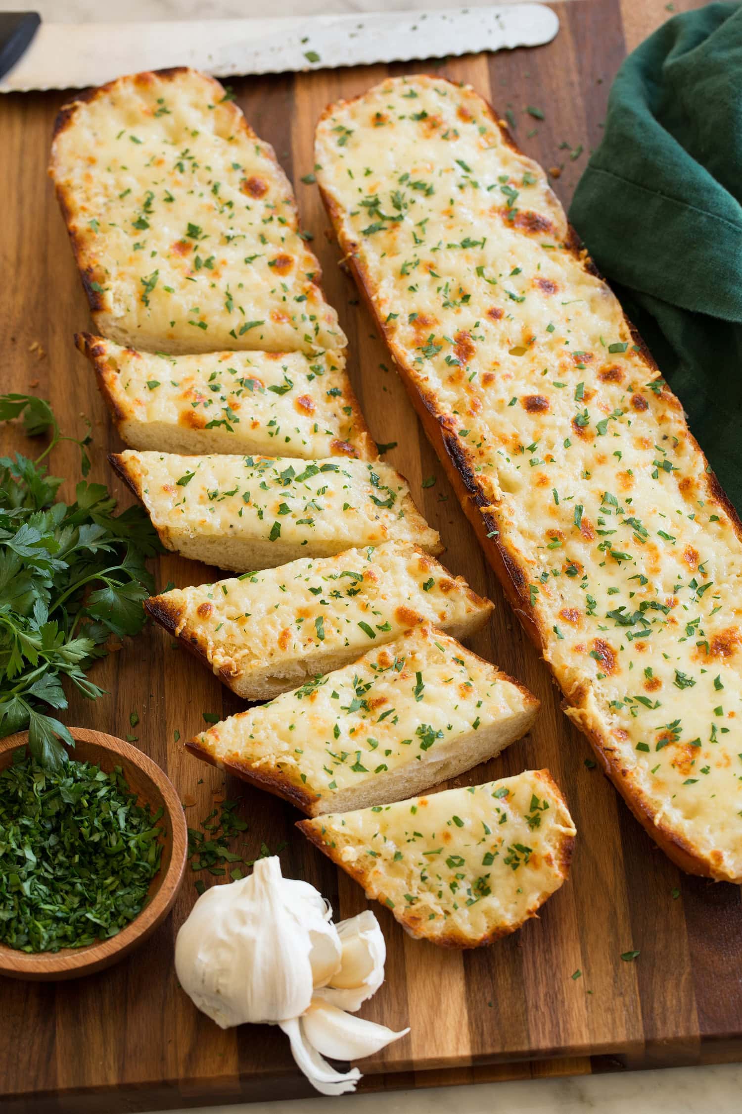 Cheesy garlic bread cut into slices with garlic and parsley shown to the side.