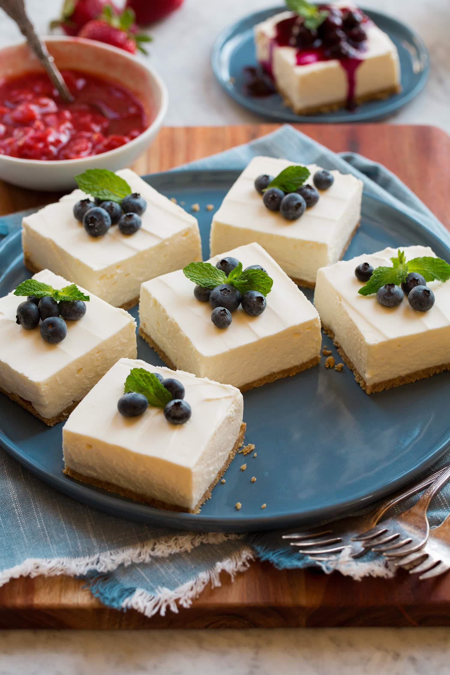No bake cheesecake shown cut into bars garnished with fresh blueberries and mint. They are resting on a large blue serving plate.