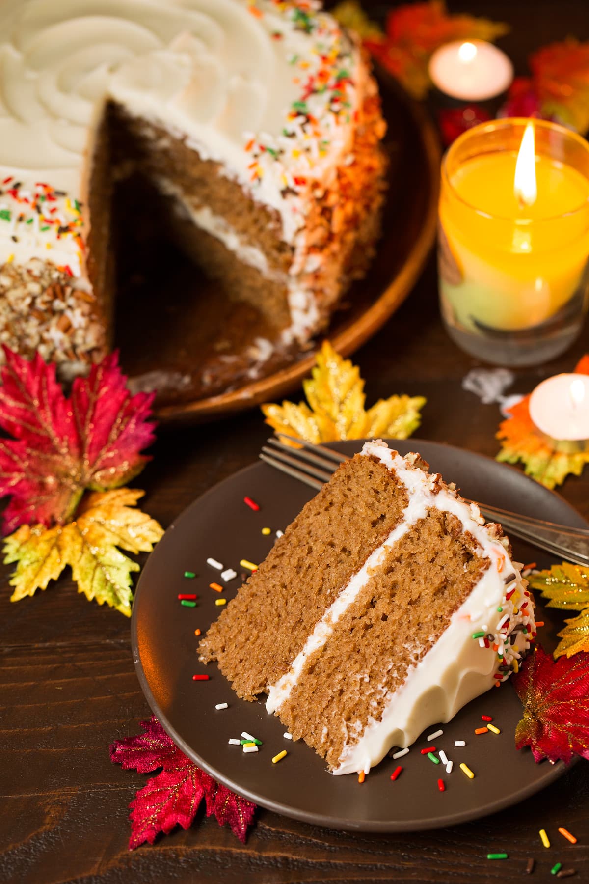 One slice of spice cake on a single serve plate and a whole spice cake is in the background. Decorations in the image include fall leaves, a lit candle and a wooden surface.