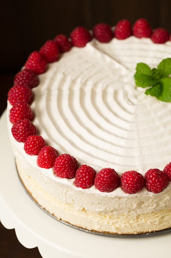 Image of a whole cheesecake decorated with swirled whipped cream on top, fresh raspberries and mint.