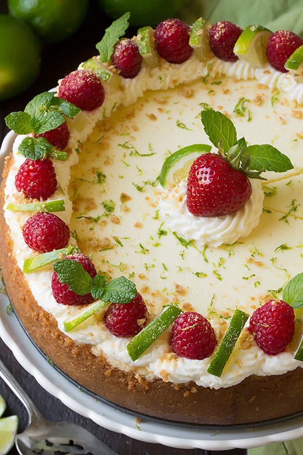 Whole Key Lime Cheesecake on a white serving plate. Cheesecake is garnished with whipped cream, raspberries, and limes along edges and a strawberry in center.