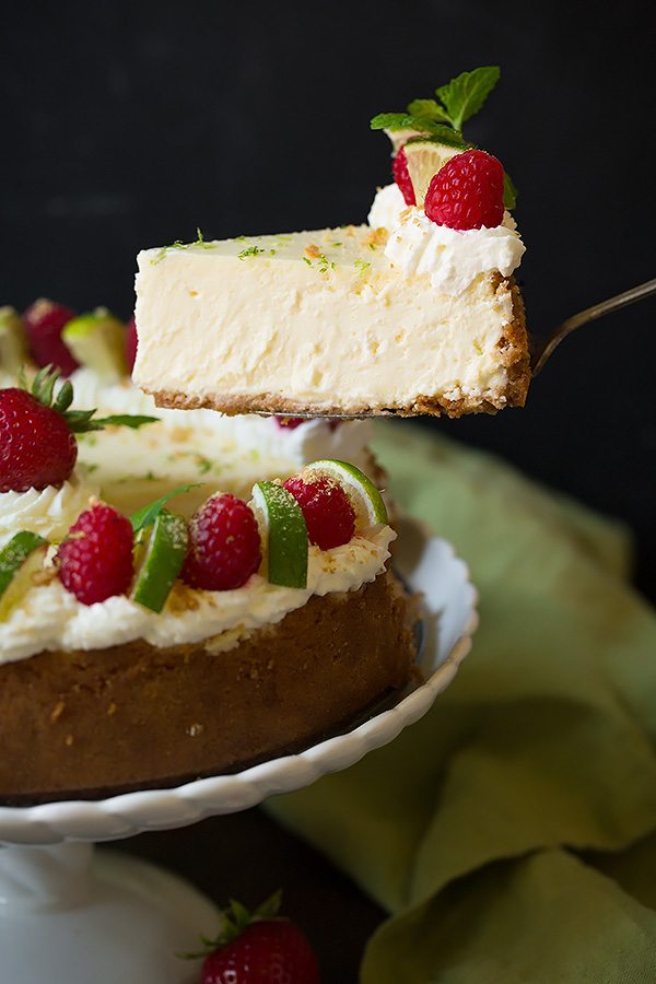 Lifting a slice of key lime cheesecake from the whole cake. It's garnished with whipped cream, raspberries, mint and graham crackers.