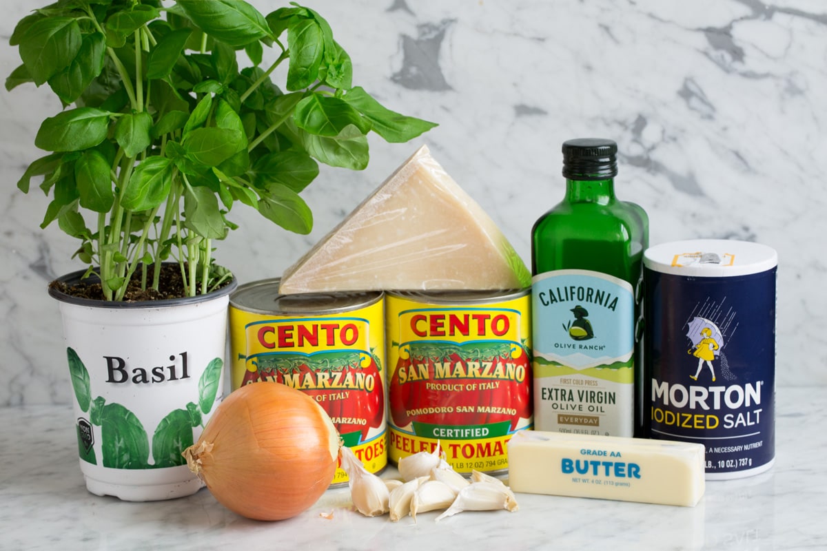 Ingredients needed to make marinara sauce shown here including canned San Marzano tomatoes, extra virgin olive oil, garlic, butter, parmesan, basil, onion, and salt.