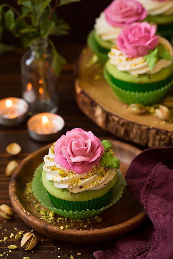 One pistachio cupcake on a wooden dessert plate. Cupcake is light green with a white buttercream frosting and it's topped with a pink piped rose.