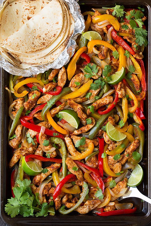 Chicken fajita mixture on a sheet pan with a side of tortillas. Fajita mixture includes seasoned chicken strips, bell peppers, onions and cilantro.