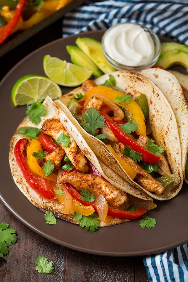 Chicken Fajitas wrapped in tortillas served with limes and sour cream on brown plate.
