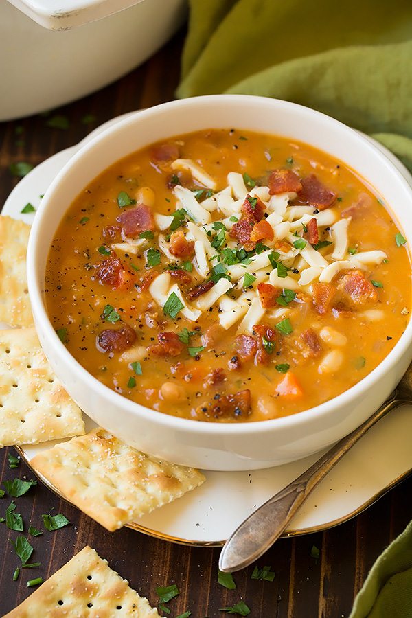 Bean and Bacon Soup in a white serving bowl set over a white plate. Soup is topped with mozzarella, bacon and parsley and served with a side of saltine crackers.