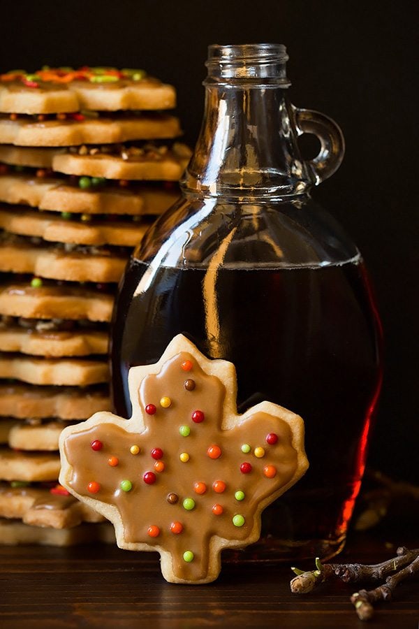 Shortbread cookie flavored with pale cut into leaf shape and set in front of a jar of maple syrup, in the background is a large stack of the shortbread cookies.