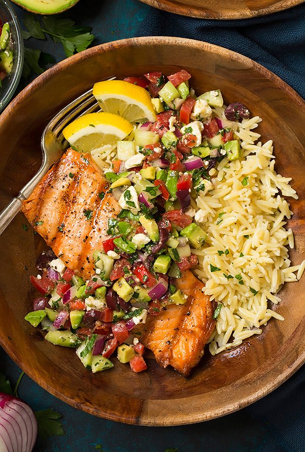 Grilled Salmon in a wooden bowl served with with Avocado Greek Salsa and a side of orzo. It's set atop a blue surface.