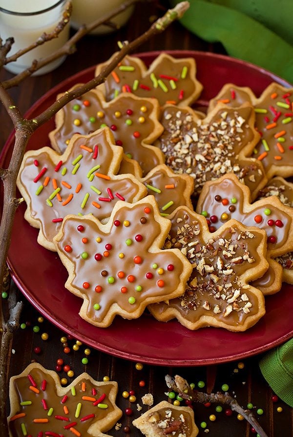 Glazed Maple Shortbread Cookies cut into leaf shapes and sitting on a maroon plate. There's a branch for decoration to the side and a green napkin.