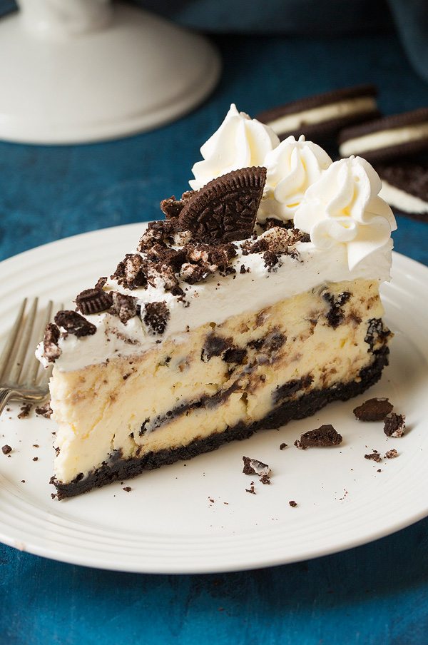 Oreo cheesecake on a white plate set over a blue surface. Cheesecake is made with Oreo crust, cream cheese and Oreo filling and topped with whipped cream.