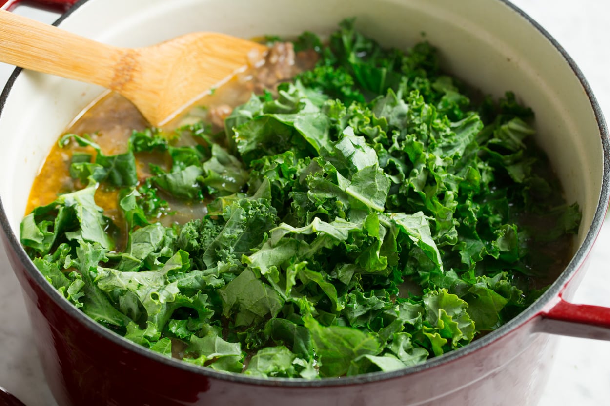Adding kale to zuppa toscana soup in pot.