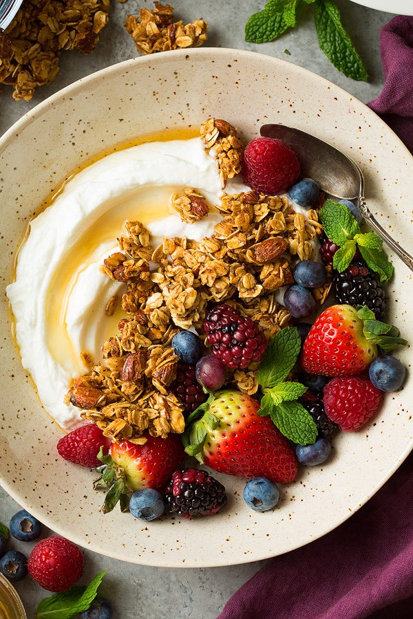 Close up image of granola, fresh berries, yogurt and honey in a speckled beige bowl.