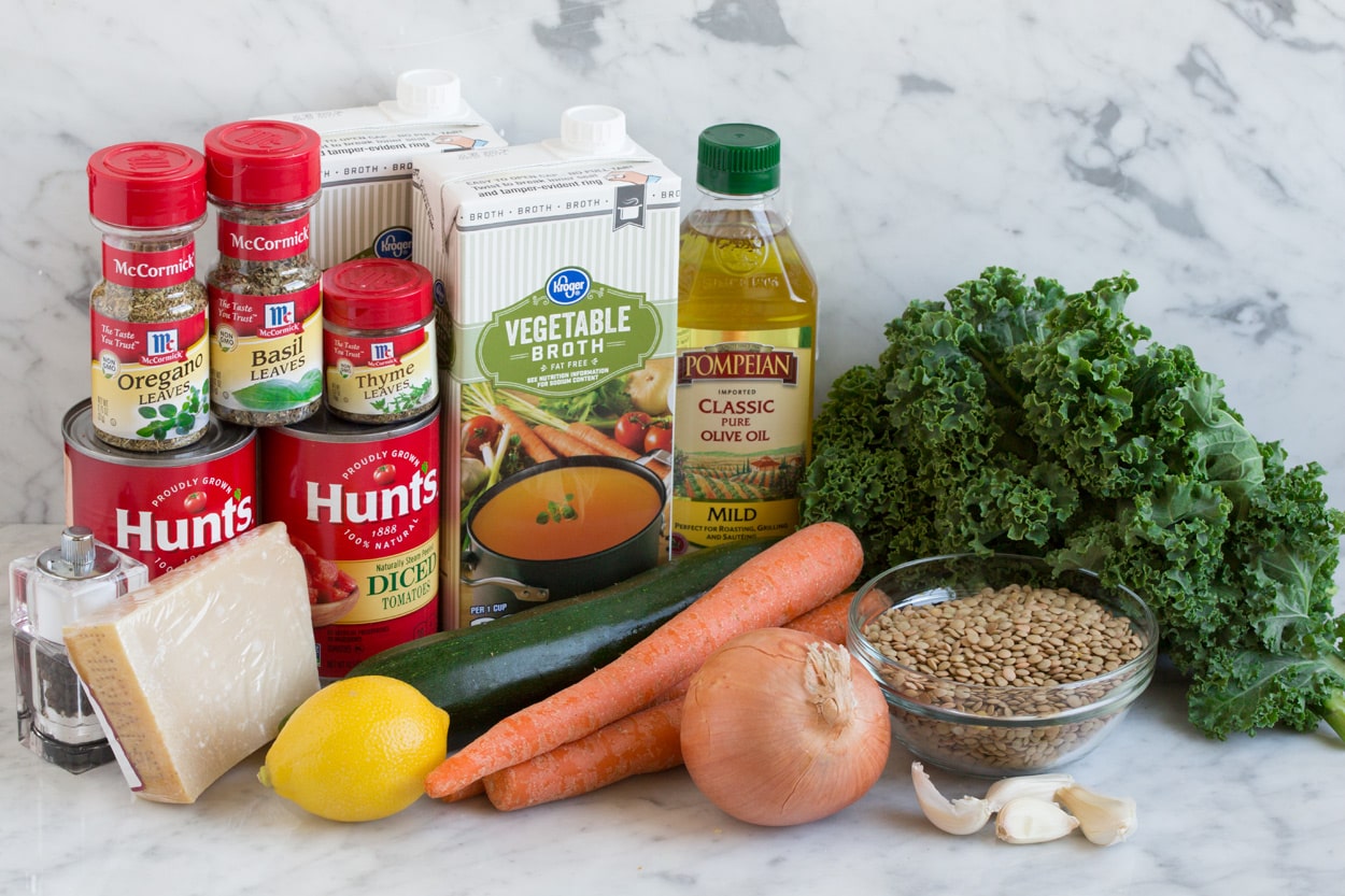 Ingredients needed to make lentil soup show n here including dry lentils, kale, onion, garlic, zucchini, lemon, vegetable broth, olive oil, canned tomatoes, dried herbs and parmesan.