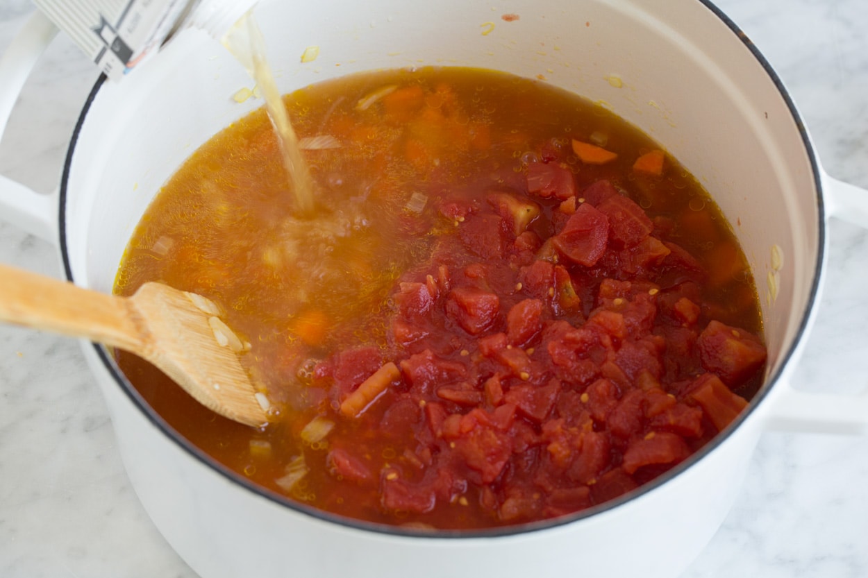 pouring broth and diced tomatoes into a large pot of lentil soup