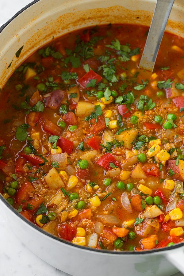 Quinoa and Vegetable Stew in a large white pot. Ingredients include potatoes, bell peppers, tomatoes, corn, peas, cilantro, broth, quinoa, spices.