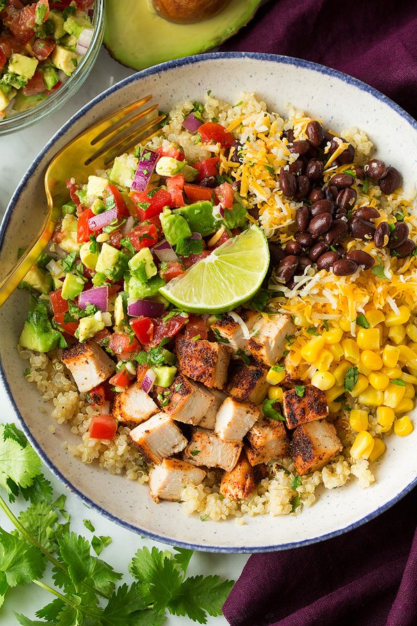 chicken Burrito bowls layered with quinoa, grilled chicken, black beans, corn, cheese and avocado salsa. Shown here in a white speckled bowl sitting on a marble surface.