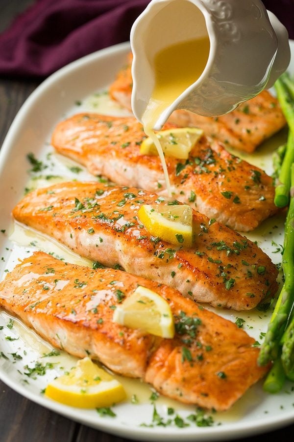 Pouring lemon butter sauce over pan fried salmon fillets on a white platter.