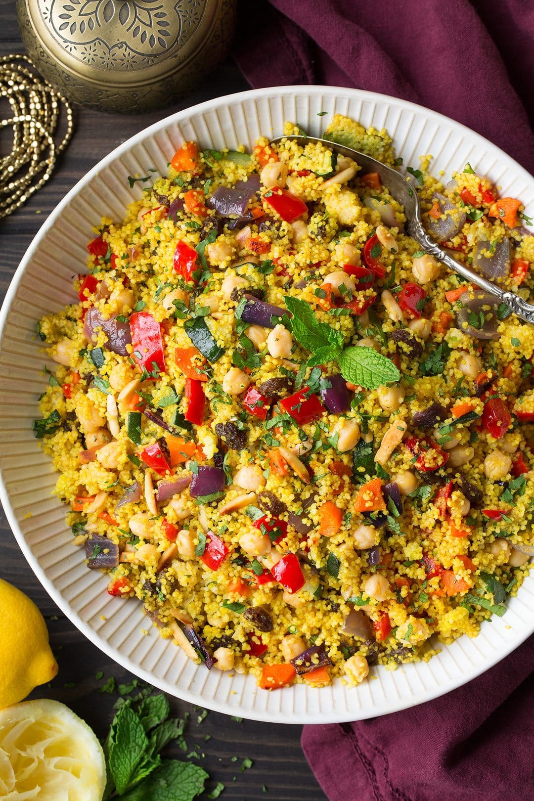 Couscous with roasted vegetables, chick peas and almonds in a large white serving bowl. Bowl is set over a dark brown wooden surface and a maroon napkin is laying to the side of the bowl.