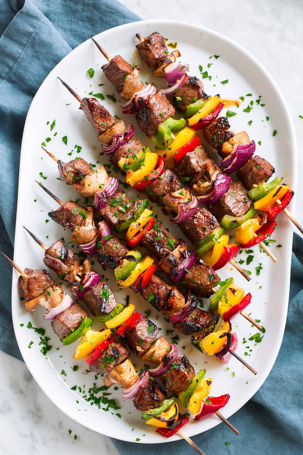 Seven steak kebabs on wooden skewers on a white platter set over a blue cloth on a marble table.
