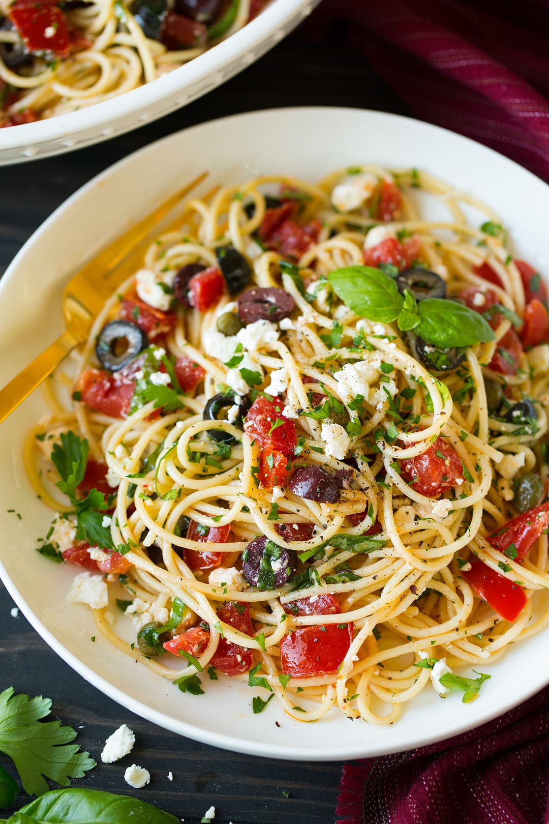 Greek spaghetti layered with tomatoes, black olives, kalamata olives, feta, basil, garlic, olive oil. Served in a pasta bowl on a brown wooden surface.