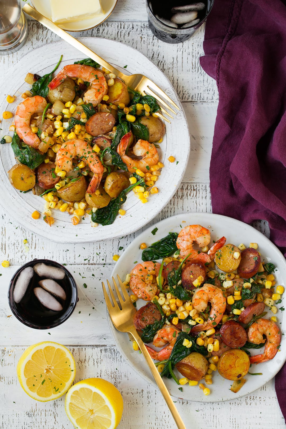 Shrimp boil shown here on two white serving plates set over a white wooden background with a maroon napkin and fresh lemons on the side.
