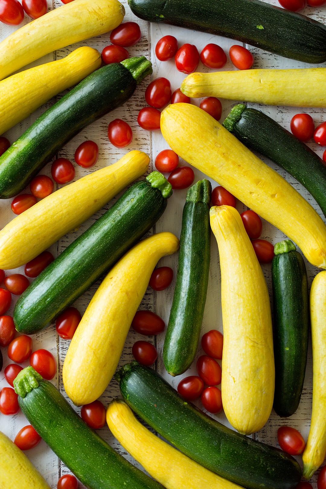 Image of fresh eight fresh zucchini, eight fresh squash and lots of fresh cherry tomatoes laying on a white wooden surface.