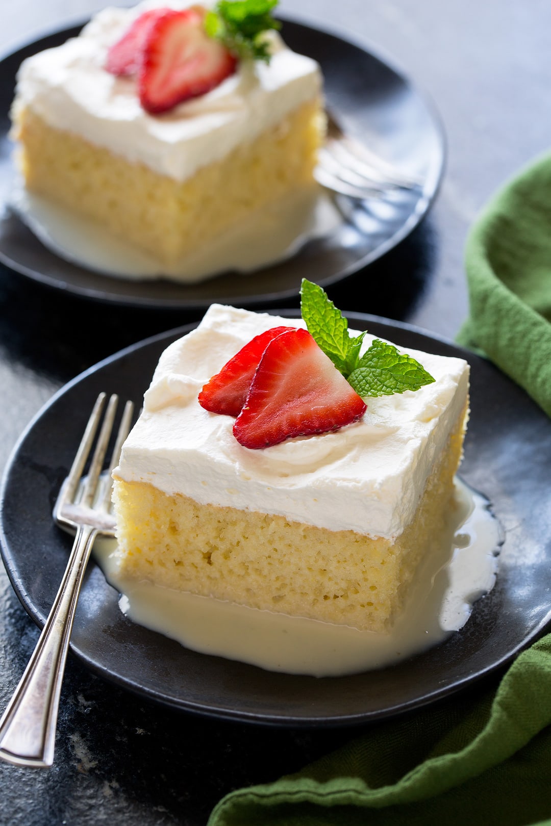 Slice of Tres Leches Cake on a small black dessert plate. Cake is a creamy yellow color with a white whipped cream frosting. It's garnished with fresh strawberry slices and mint on top.