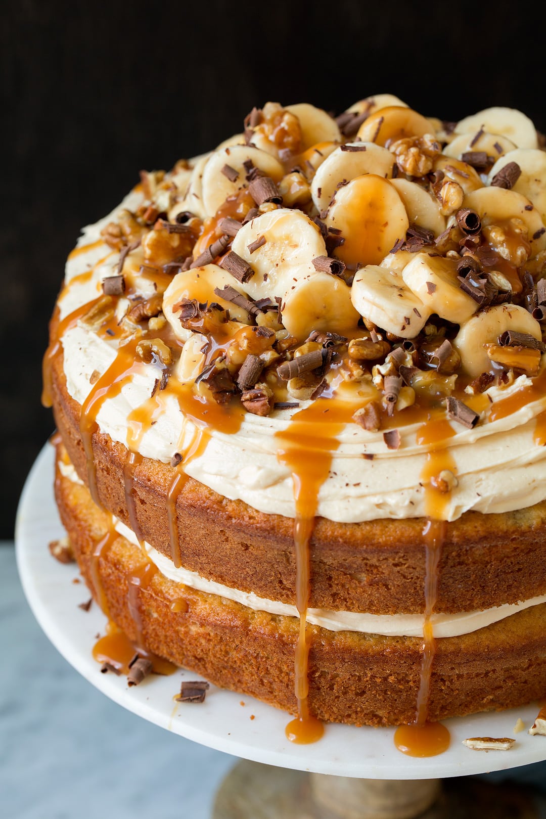 Banana cake on a cake stand. Cake is layered with cream cheese, caramel sauce, walnuts, banana slices and chocolate shavings. 