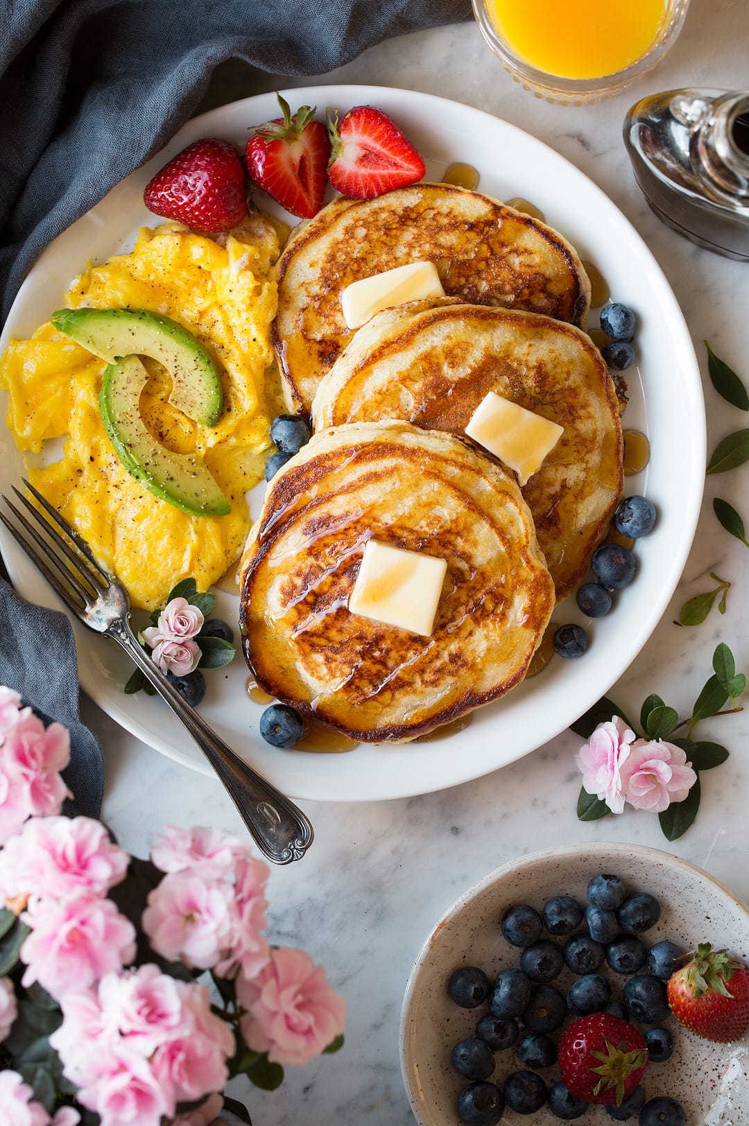 Three buttermilk pancakes on a large white plate dotted with slabs of butter and drizzled with maple syrup. Included on the side are scrambled eggs and fresh fruit. 